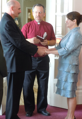 wedding officient in red shirt and white collar presiding over grrom in black and bride in blue